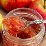 Glas med hjemmelavet æbleskrælmarmelade, med en fin teske, omgivet af friske røde æbler med skræl på et skærebræt.