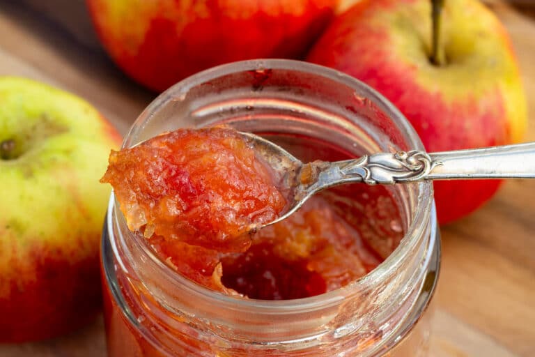 Glas med hjemmelavet æbleskrælmarmelade, med en fin teske, omgivet af friske røde æbler med skræl på et skærebræt.