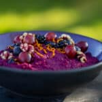 Skål på grøn baggrund med lilla grød af rødkål, gulerødder og aroniabær, toppet med valnødder, blåbær, brombær, druer og appelsinskal