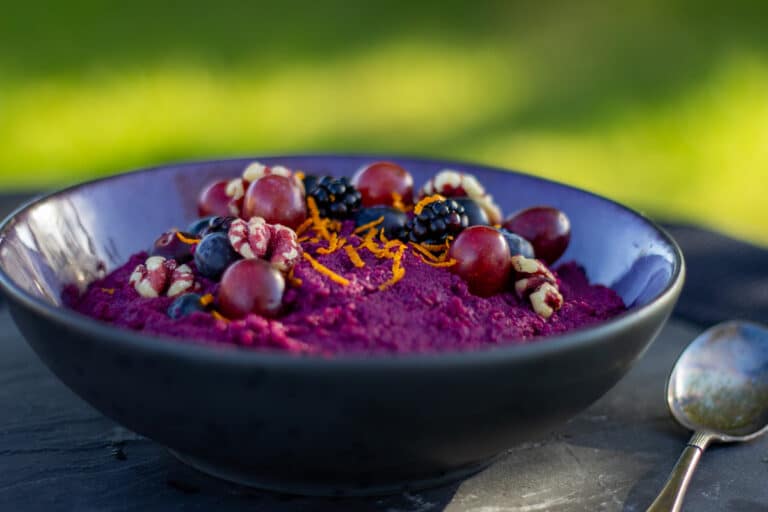 Skål på grøn baggrund med lilla grød af rødkål, gulerødder og aroniabær, toppet med valnødder, blåbær, brombær, druer og appelsinskal