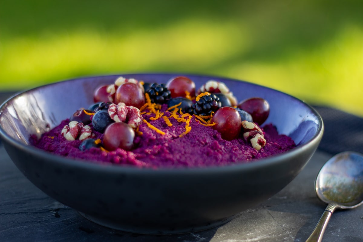 Skål på grøn baggrund med lilla grød af rødkål, gulerødder og aroniabær, toppet med valnødder, blåbær, brombær, druer og appelsinskal