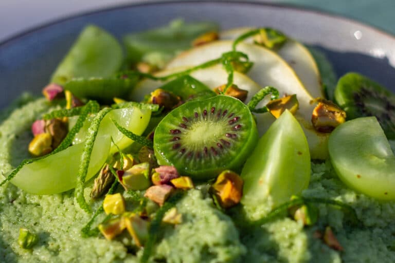 Lysegrøn energigrød pyntet med kiwibær, grønne dryer, pære, limeskal og pistcienødder