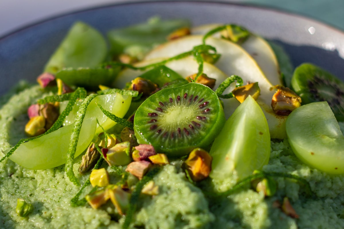 Lysegrøn energigrød pyntet med kiwibær, grønne dryer, pære, limeskal og pistcienødder