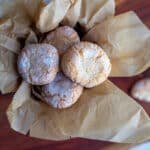 Amaretti mandelkager rullet i flormelis i en dåse foret med bagepapir og en skål med mandler ved siden af.