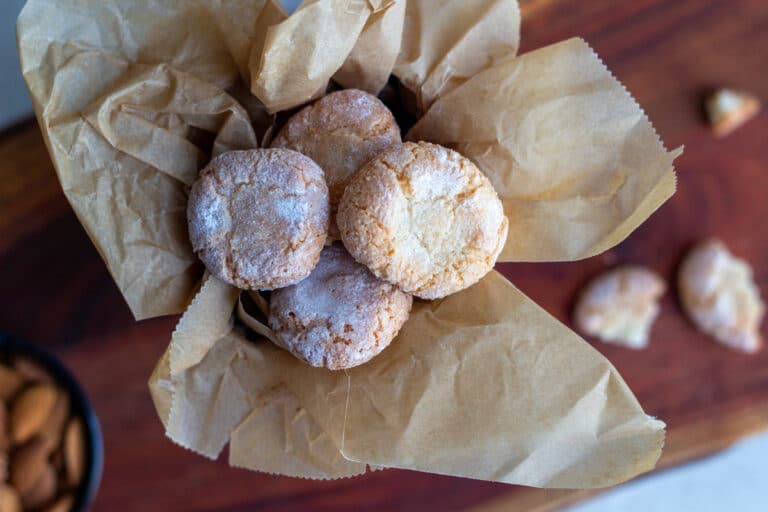 Amaretti mandelkager rullet i flormelis i en dåse foret med bagepapir og en skål med mandler ved siden af.